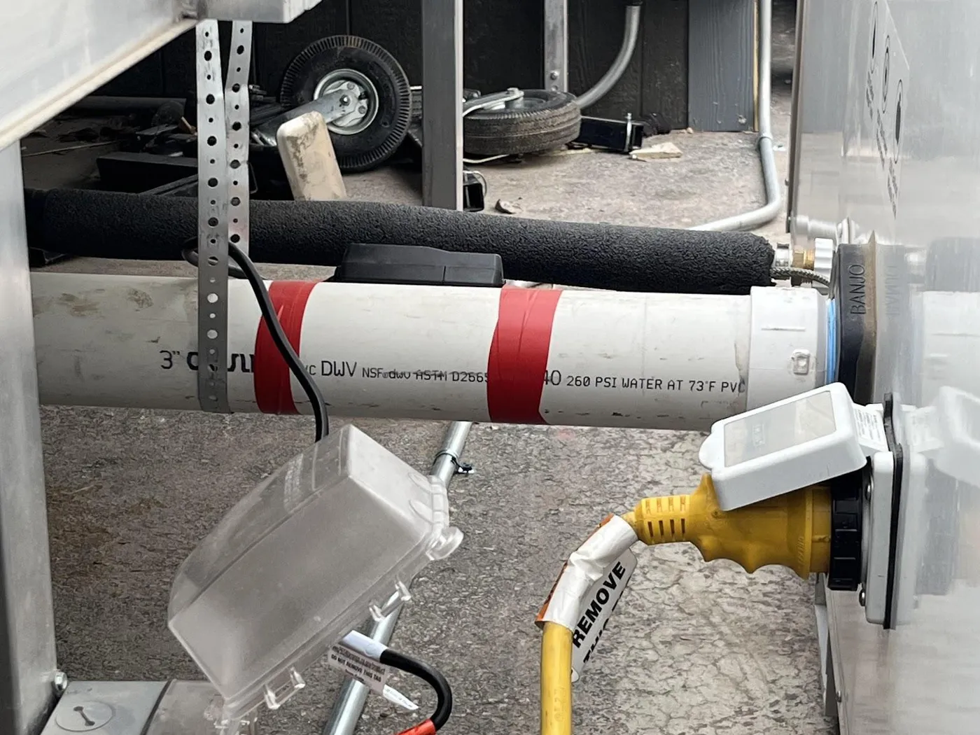 Installed connections between an office trailer and the box: insulated fresh water line, 3-inch waste line with heat cable, and 30-amp power cord to a covered outlet